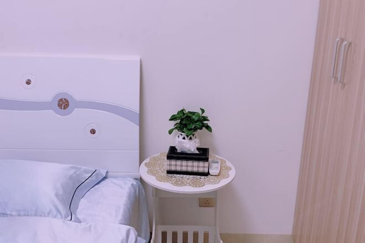A bedroom with a bed, a bedside table with a plant, a book, and a tissue box.