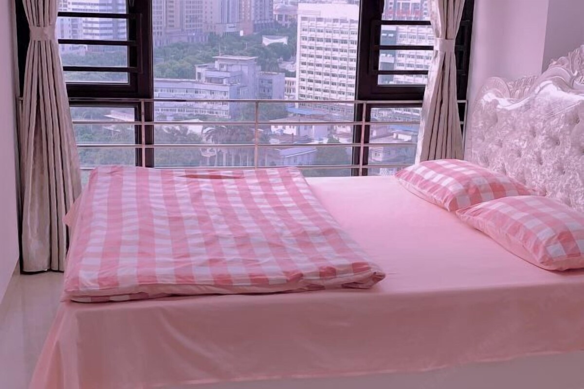 A bed with a pink checkered bedspread and matching pillows, a white upholstered headboard, and a city view through large windows.