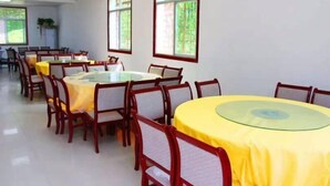 A dining area with round tables and chairs, large windows with green foliage, and a clean, white floor.