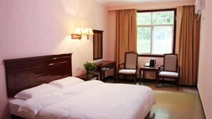 A hotel room with a bed, a desk, two chairs, a window with curtains, and a wall-mounted lamp.