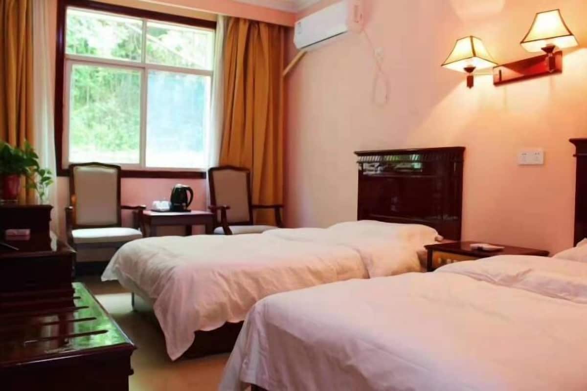A hotel room with two beds, a piano, a small table with a kettle, and a view of greenery through the window.