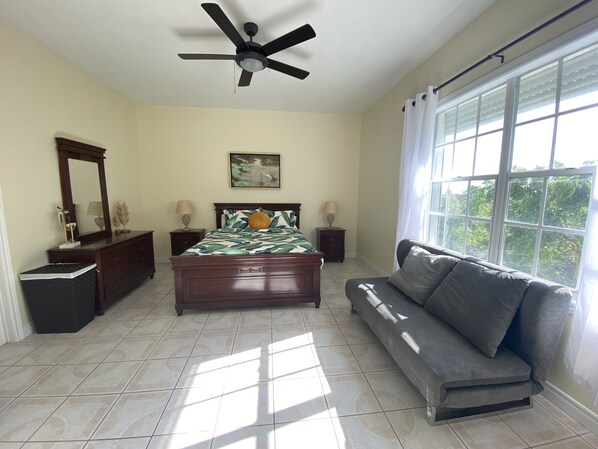 A bedroom with a bed, a nightstand, a dresser, a ceiling fan, a window with curtains, and a view of trees. - Bliss 101: Poolside Condo w/ Wraparound Balcony & Ocean View (The Bight Settlement)