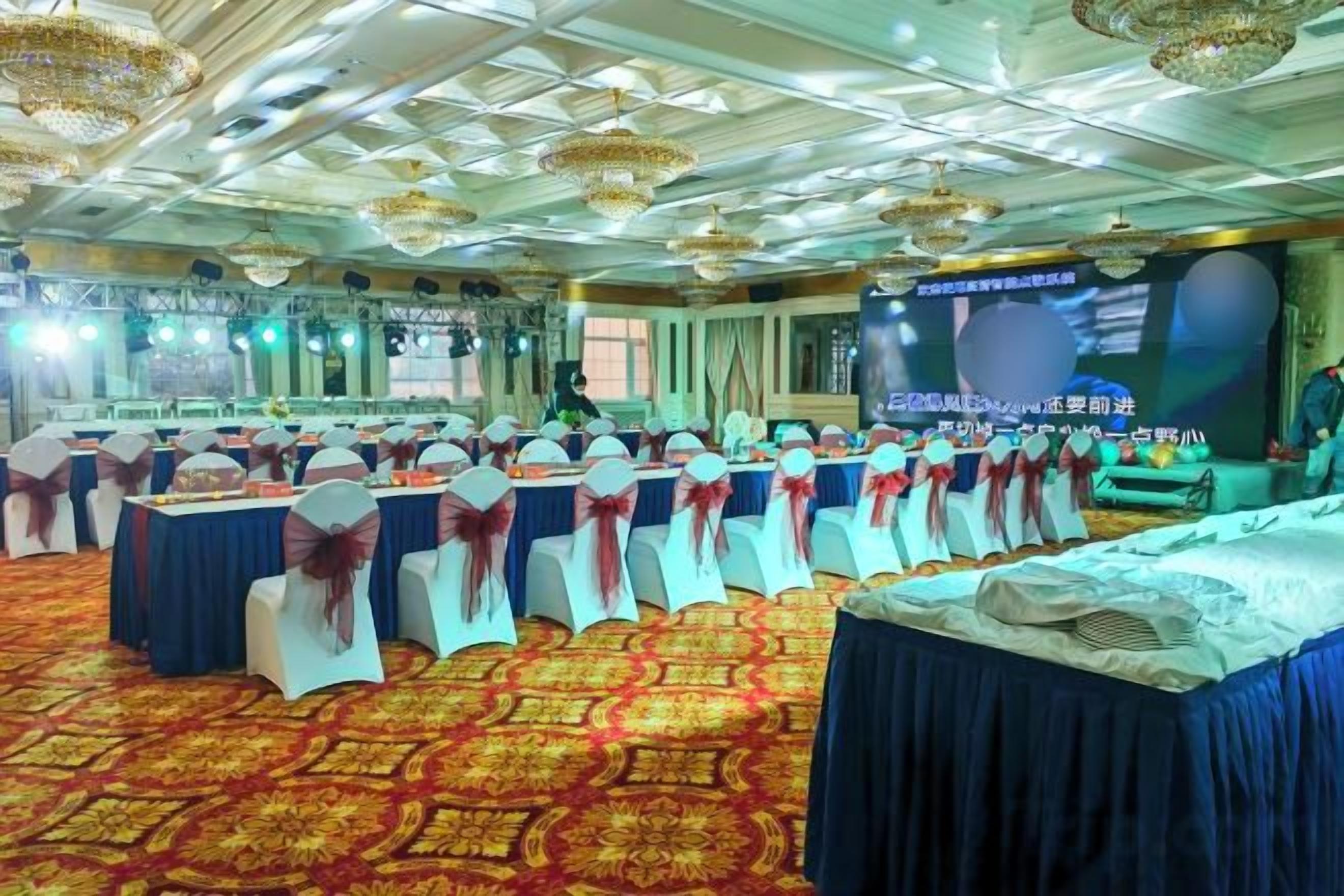 Un salón de banquetes con mesas redondas, sillas blancas y un escenario.