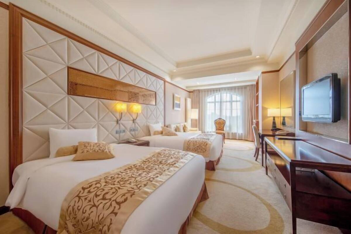 A hotel room with two beds, a desk, a TV, and a large window.