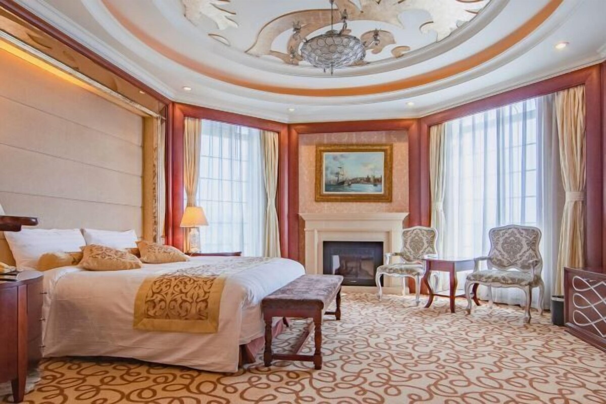 A hotel room with a large bed, a fireplace, two armchairs, and a patterned carpet.