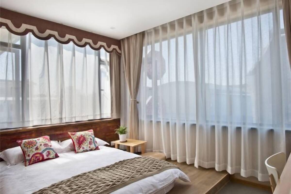 Une chambre moderne avec un grand lit, une tête de lit en bois, une petite table de chevet agrémentée d’une plante et des rideaux légers.
