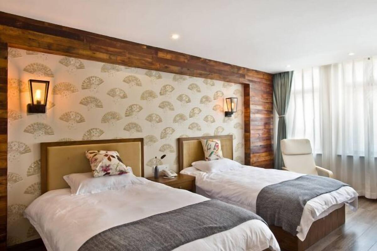 Une chambre d’hôtel avec deux lits, des têtes de lit en bois, un papier peint à motifs et une chaise près de la fenêtre.