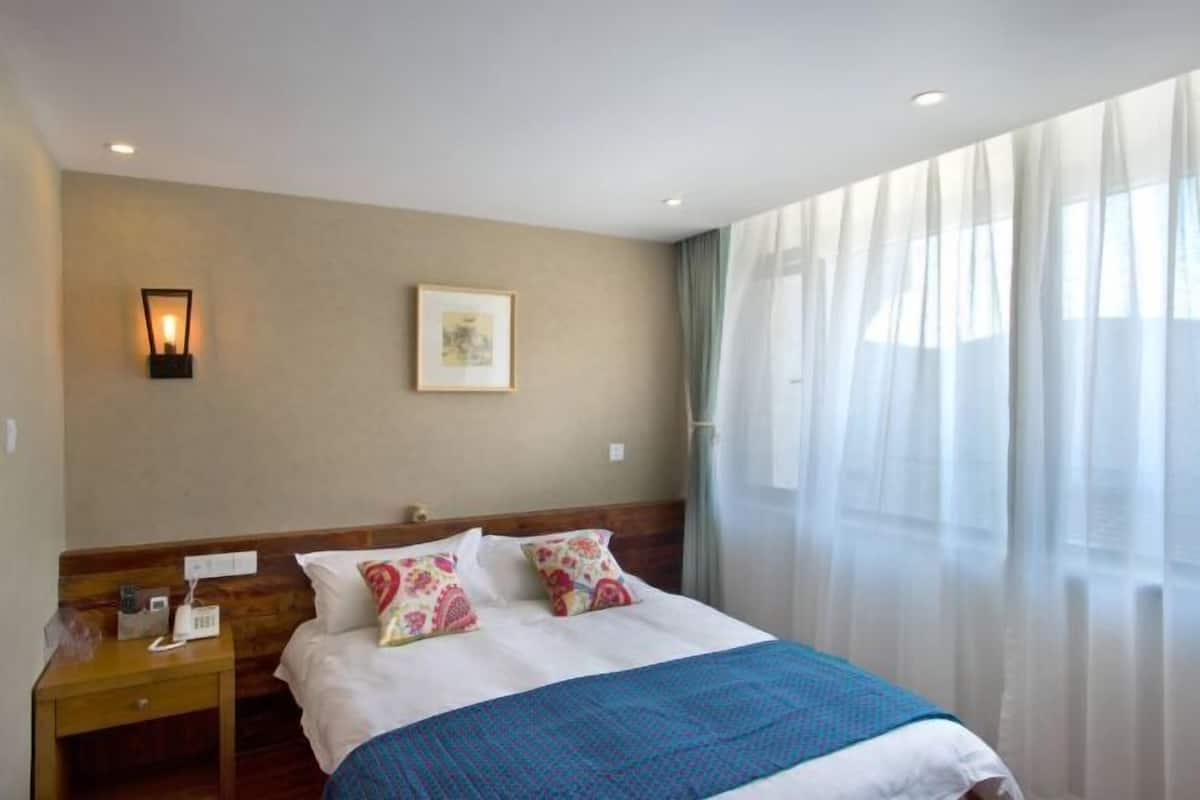 Une chambre d’hôtel avec un lit, une table de chevet, une lampe fixée au mur, un tableau encadré et des rideaux légers.