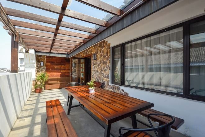 A covered outdoor area with a wooden table and chairs, stone walls, and a large window.