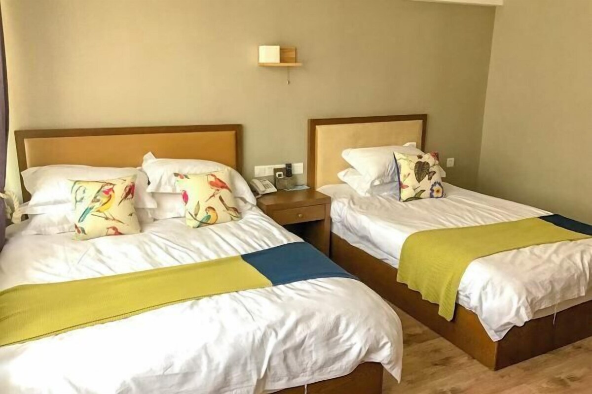 Two twin beds with white bedding and colorful pillows, a bedside table with a lamp, and a wall-mounted light fixture.