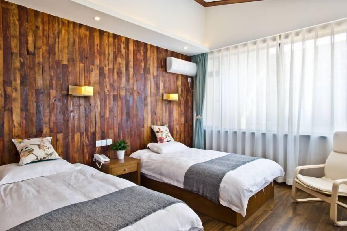 A hotel room with two beds, wooden panel walls, a modern chair, and a large window with curtains.