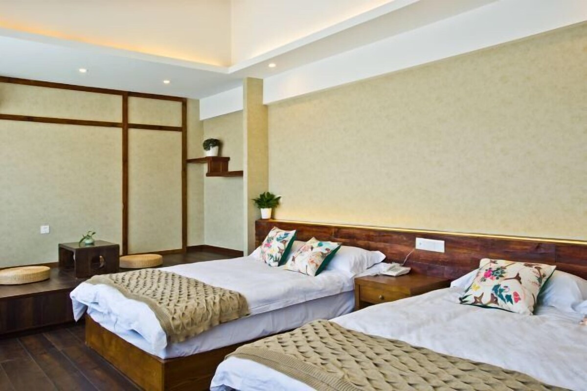 A hotel room with two beds, a small table, a shelf with a plant, and a wooden headboard.