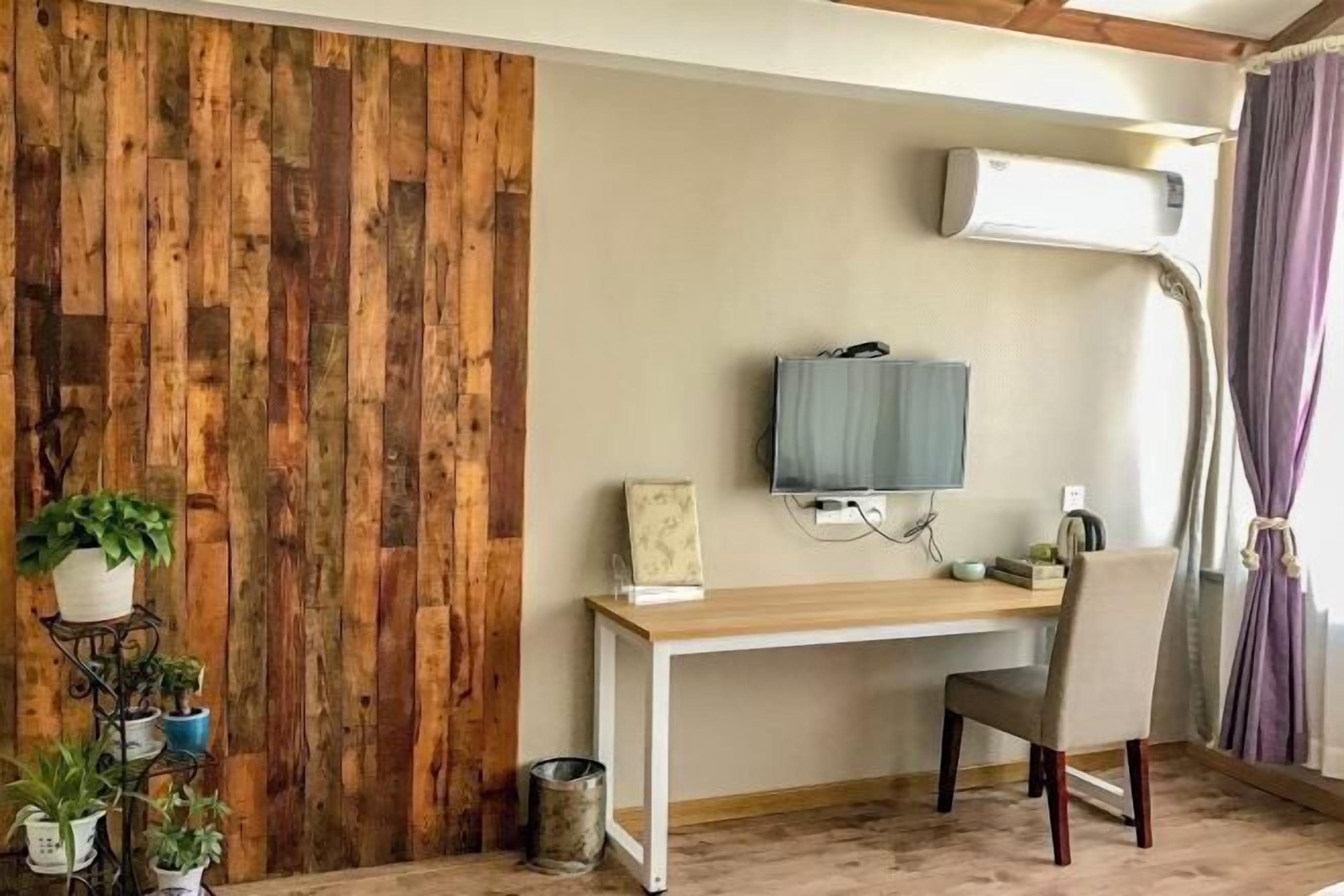 A room with a wooden wall panel, a desk with a chair, a mounted TV, and a window with curtains.