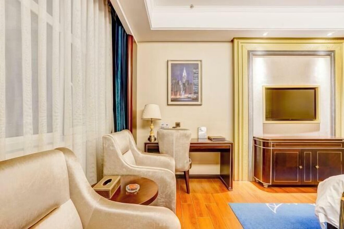A hotel room with a large bed, two armchairs, a small table, a desk, a television, and a framed picture on the wall.