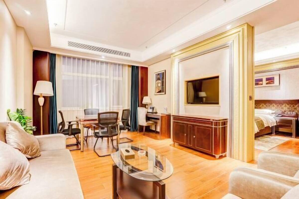 Quarto de hotel com sofá, mesa de centro de vidro, escrivaninha com cadeiras, televisão, cama e guarda-roupa.