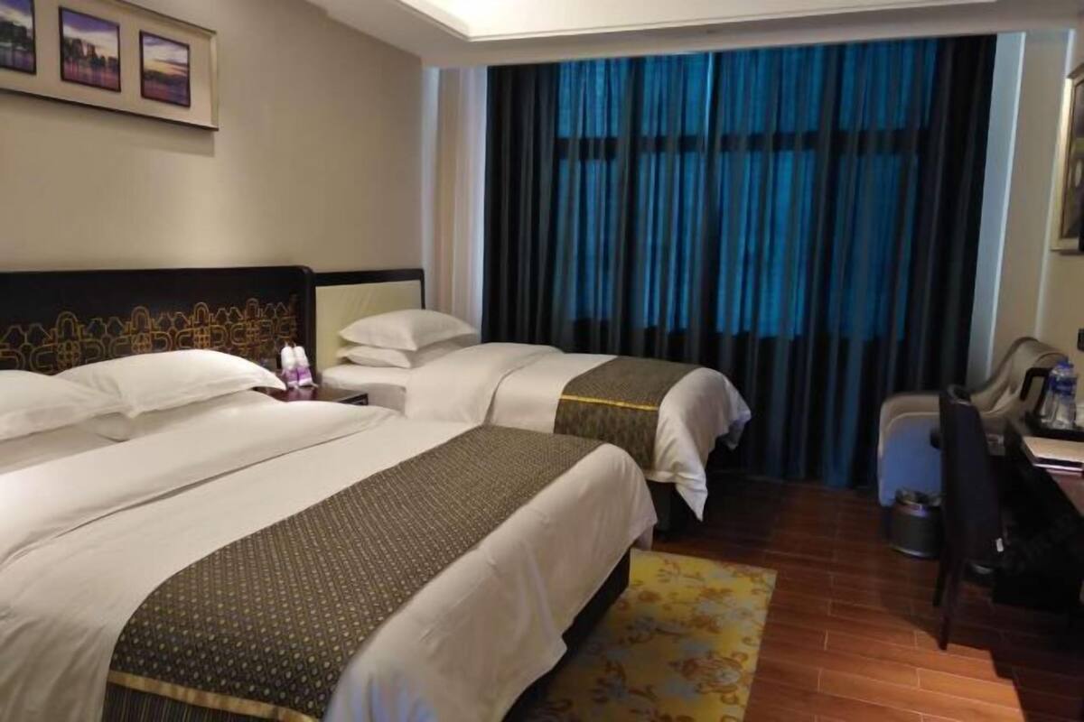 Quarto de hotel com duas camas, uma escrivaninha, uma cadeira e vista para a cidade através das cortinas.