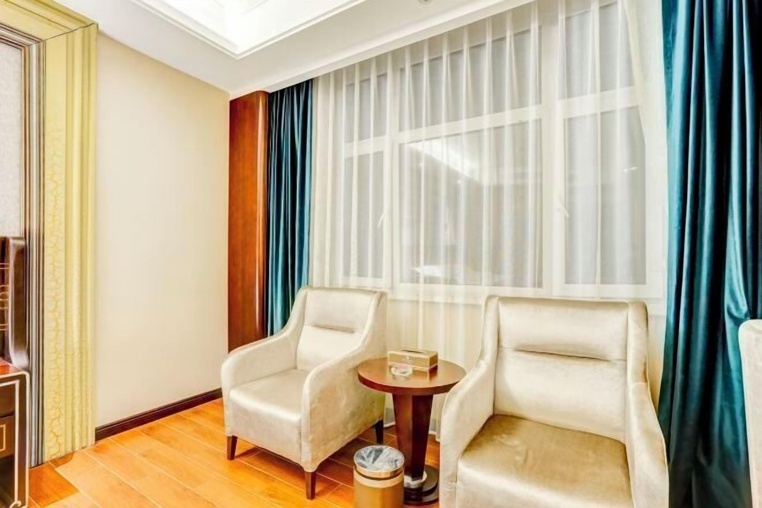 Habitación de hotel con dos sillones color beige, una mesita redonda y una ventana con cortinas blancas traslúcidas y un cortinero azul.