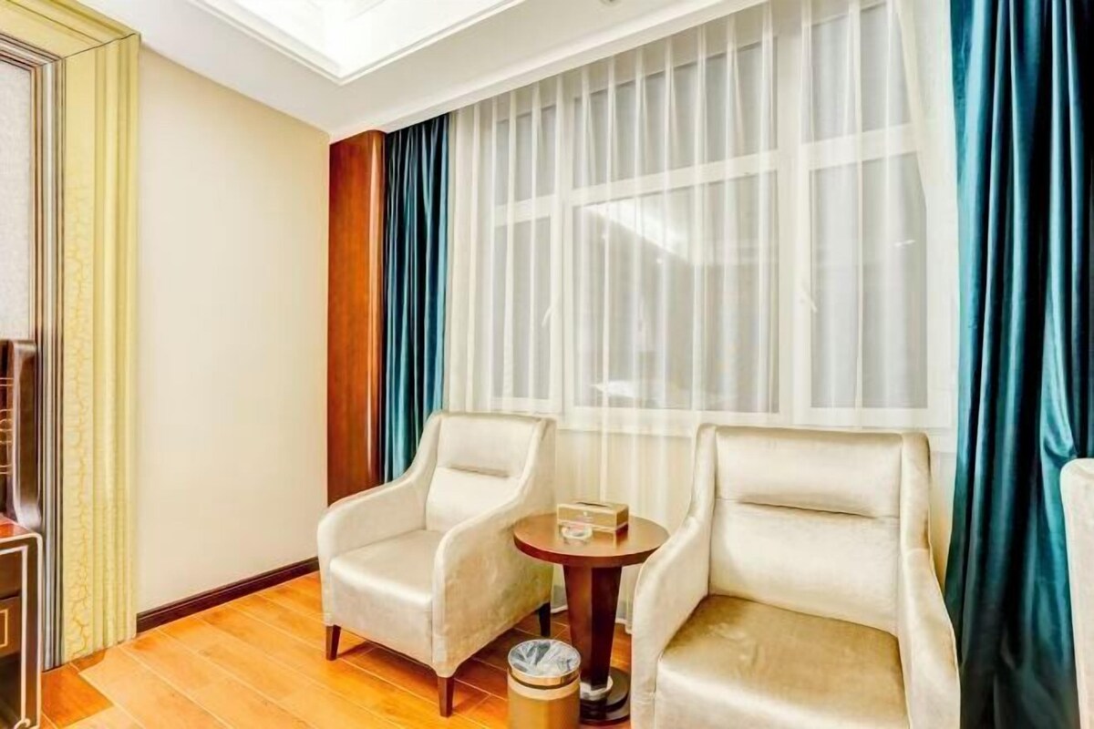 A hotel room with two beige armchairs, a small round table, and a window with sheer white curtains and a blue drape.
