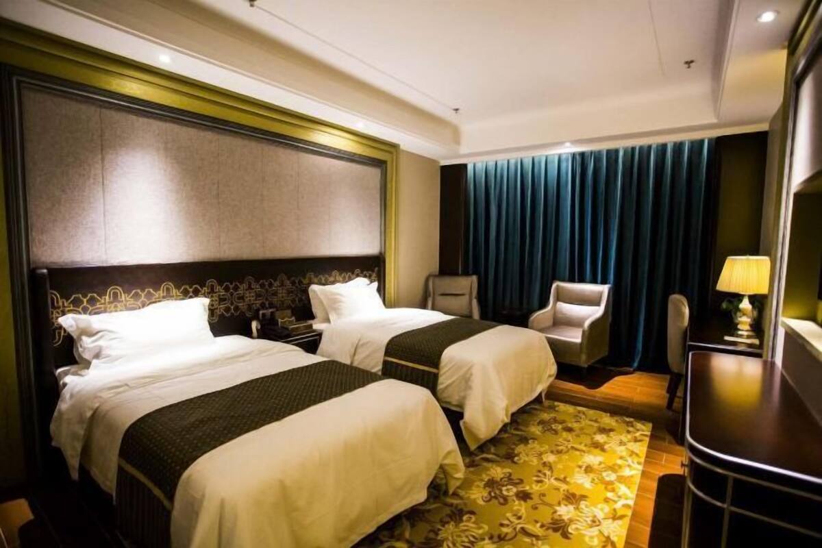 Quarto de hotel com duas camas, uma escrivaninha, uma cadeira e uma janela grande com cortinas.