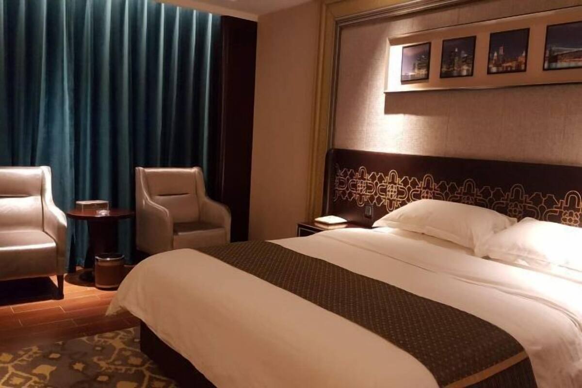 Quarto de hotel com uma cama grande, duas poltronas, uma mesa lateral e vista para a cidade pela janela.