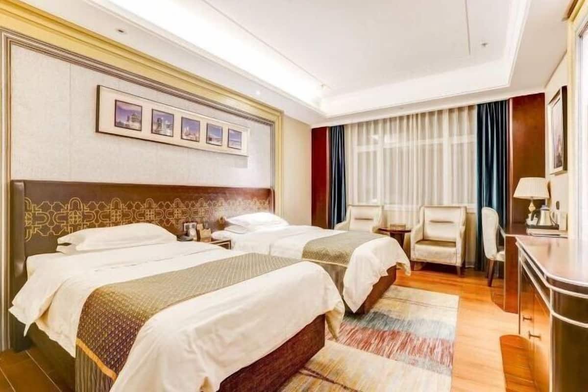 Quarto de hotel com duas camas, uma escrivaninha, uma cadeira e uma janela grande com cortinas.