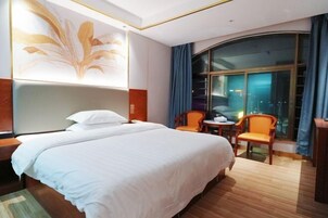 Habitación de hotel con una cama grande, dos sillas, una mesita, un ventanal con cortinas y una obra de arte colgada en la pared.