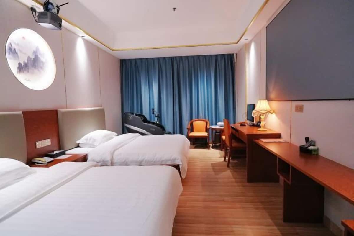 A hotel room with two beds, a desk, a chair, a lamp, and a projector mounted on the ceiling.