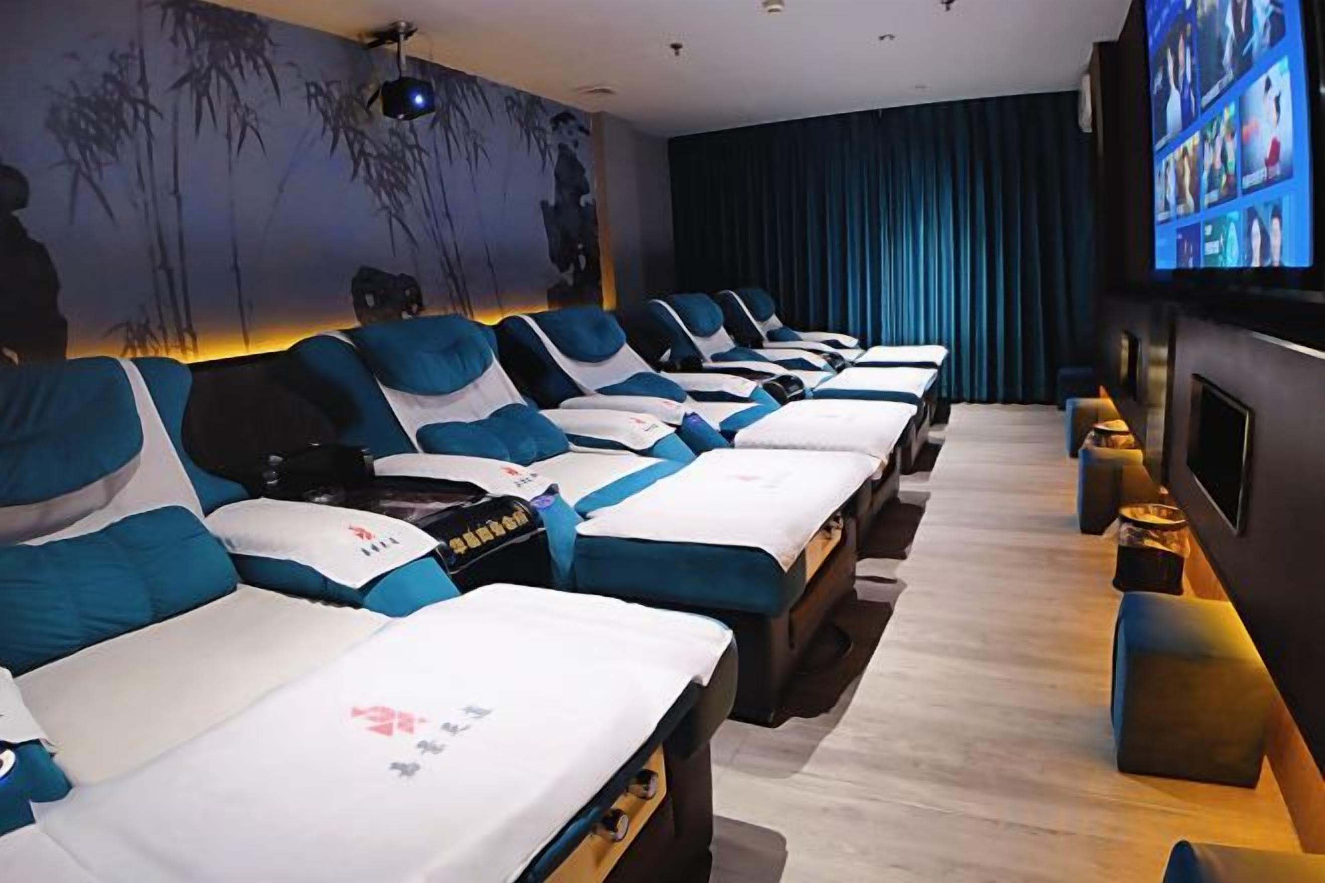 A row of massage beds with blue and white linens, a wall-mounted TV, and a dark curtain backdrop.