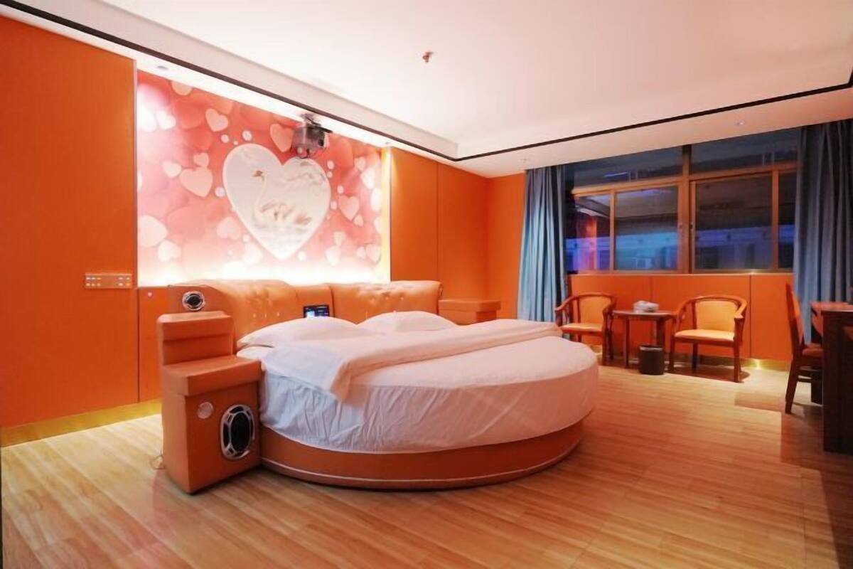 A circular bed in a room with orange walls, a heart-themed wall decoration, and a view of the city through a window.