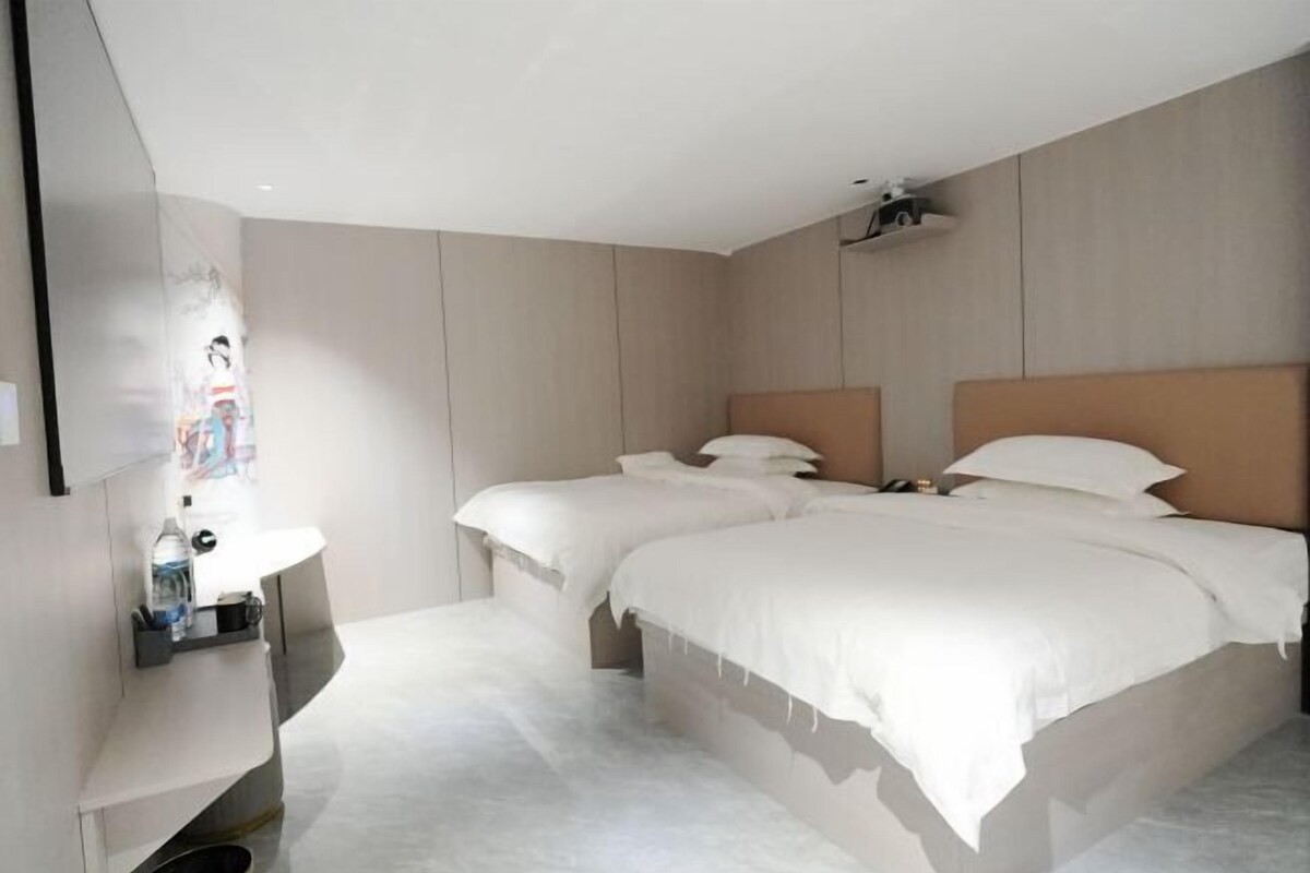A modern hotel room with two beds, a built-in TV, and a small bedside table with a bottle and glasses.