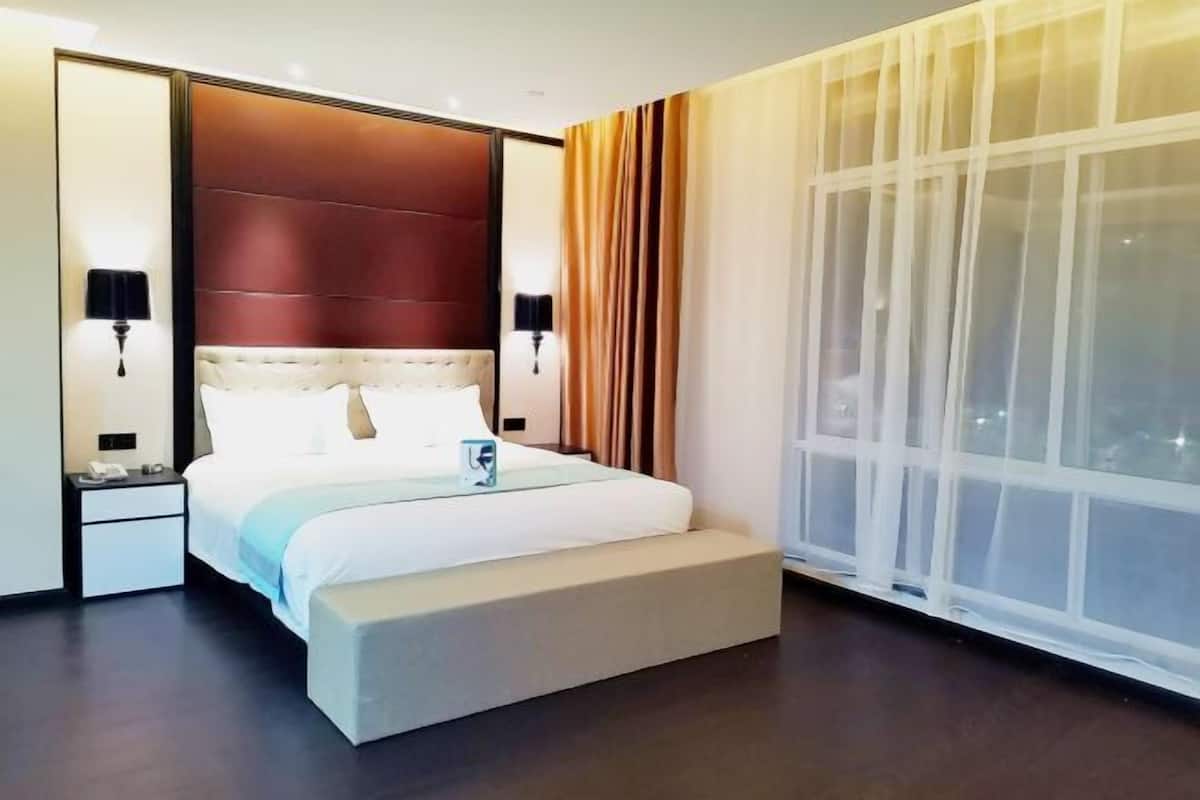 Una habitación de hotel moderna con una cama grande, una mesita de noche, una lámpara de noche y un amplio ventanal con cortinas traslúcidas.