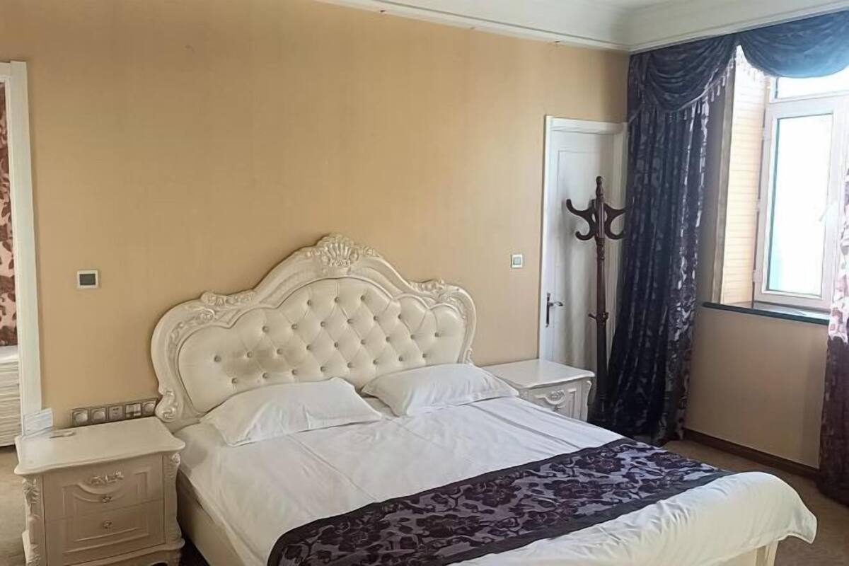 Une chambre à coucher avec un lit doté d’une tête de lit capitonnée, une table de chevet, une armoire et une fenêtre avec des rideaux.