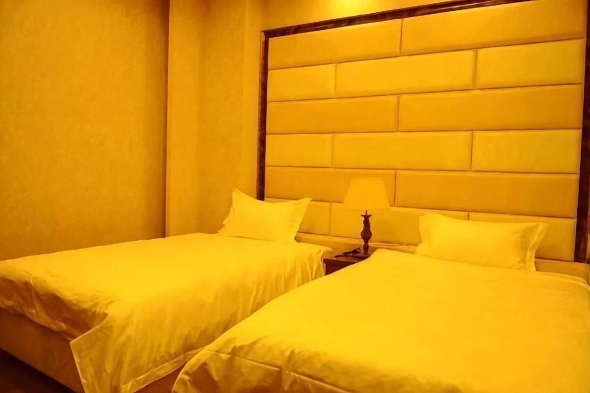 Quarto de hotel com duas camas, cabeceira e mesa de cabeceira com abajur.