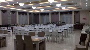 A large banquet hall with multiple tables and chairs, ceiling lights, and a stage area.