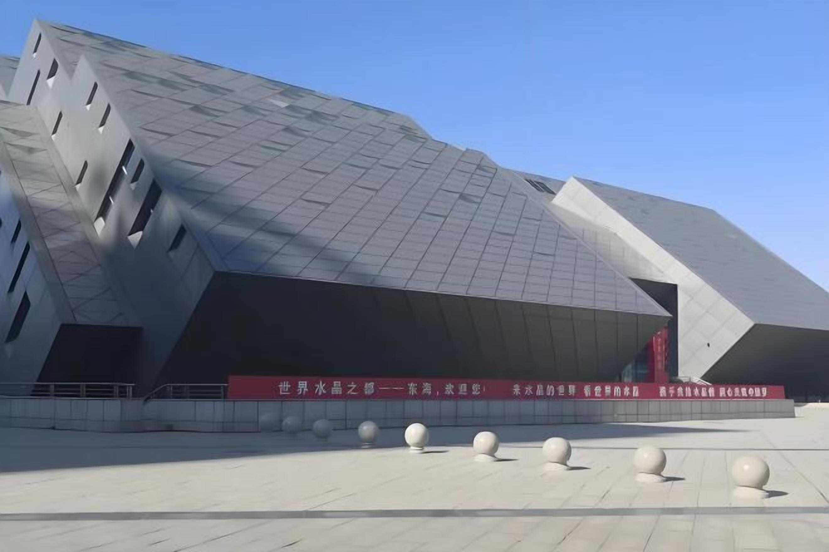Un edificio moderno dal design angolare particolare, con un'ampia piazza aperta, dotata di dissuasori sferici e uno stendardo rosso con testo bianco.