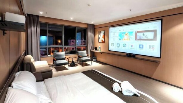 A modern hotel room with a large bed, a seating area with a table, and a large screen displaying a presentation. - China Inn (Donghai Xingfu Bridge) (Donghai)