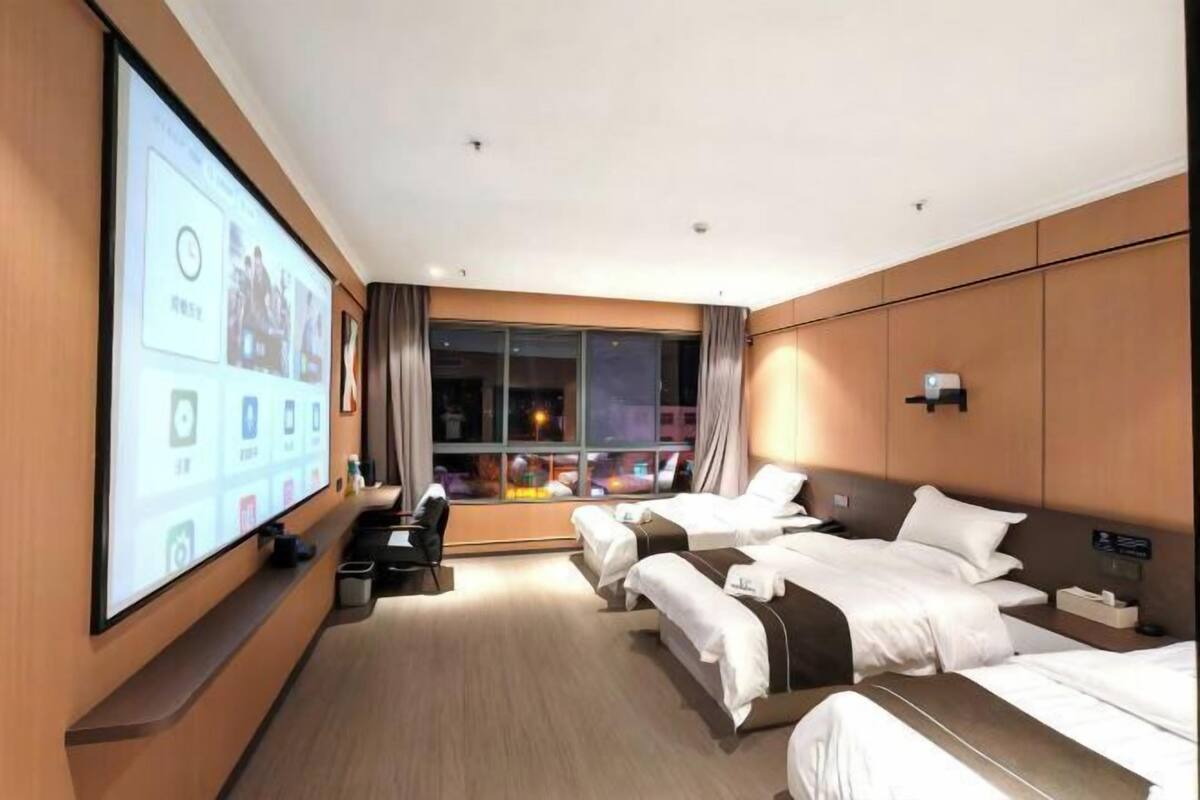 A hotel room with two beds, a large screen displaying a digital interface, a desk with a chair, and a window with curtains.
