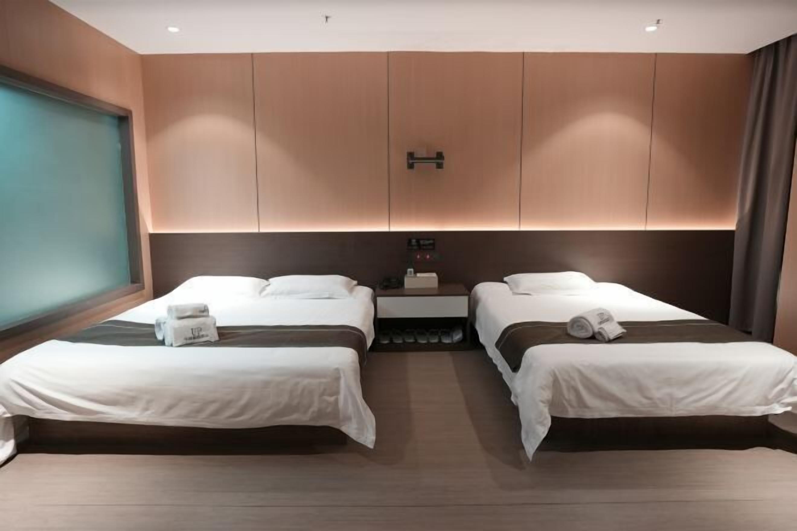 Une chambre d’hôtel avec deux lits, une grande fenêtre, un panneau de commande fixé au mur et une petite table avec une lampe.