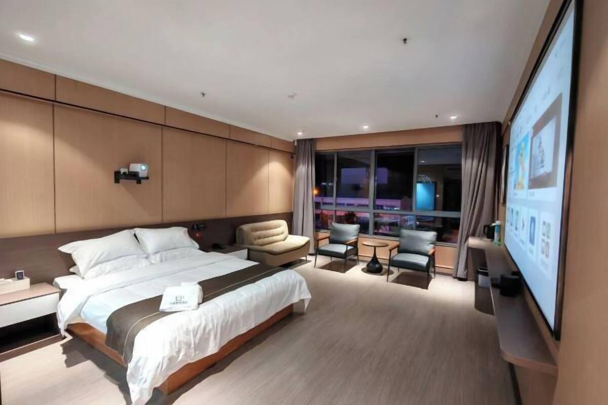 A modern hotel room with a large bed, a seating area with a sofa and chairs, a small table, and a large window with curtains.