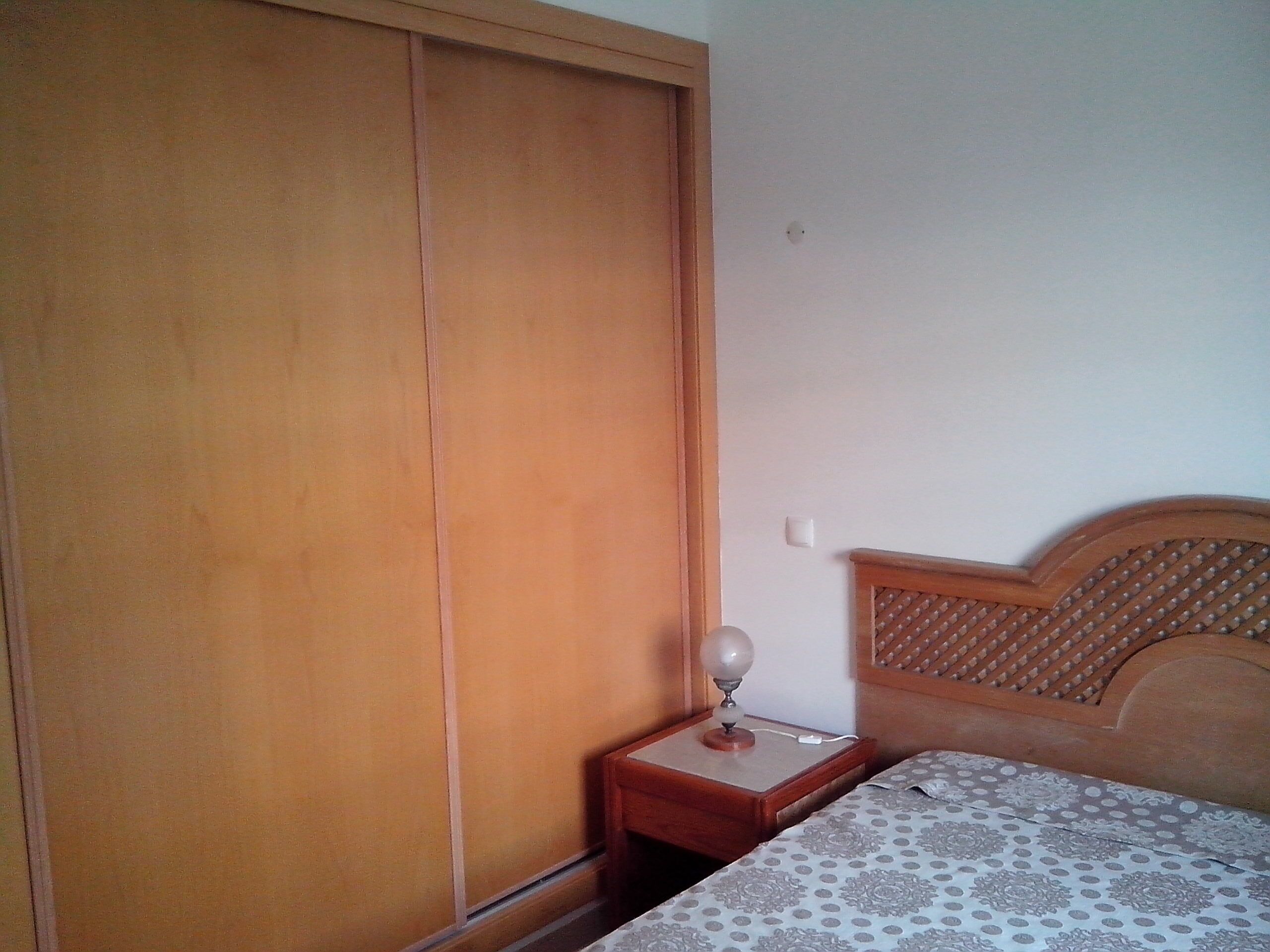Een slaapkamer met een houten garderobe, een nachtkastje met een lamp en een bed met een decoratieve spreideken.