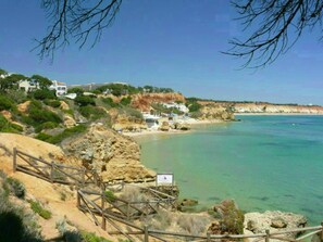 Une zone côtière avec une plage de sable, des falaises rocheuses et une mer bleue et limpide. On aperçoit des bâtiments blancs sur les falaises et une clôture en bois au premier plan.