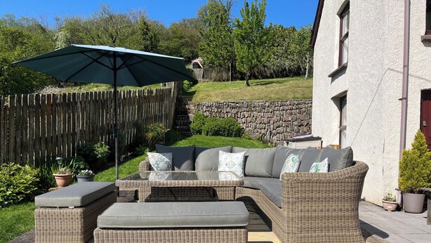 An outdoor patio area with wicker furniture, including a sofa and ottomans, and a large umbrella providing shade. - 1 Bed House - Garden, Parking, Pets Allowed (Lynton)