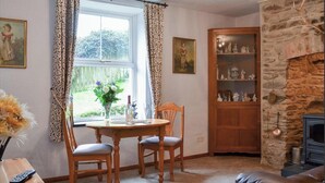 A cozy room with a stone fireplace, a wooden cabinet with figurines, and a dining area with a table and chairs. - 1 Bed House - Garden, Parking, Pets Allowed (Lynton)