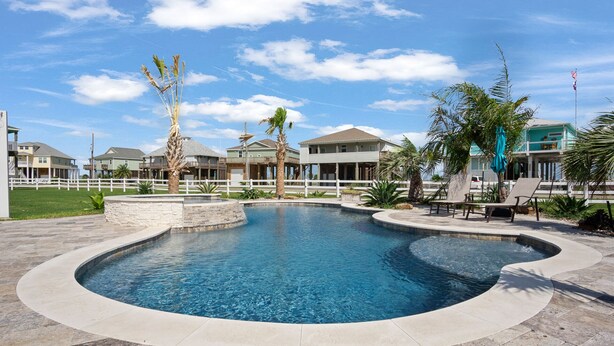 A swimming pool with a curved edge, surrounded by a stone deck. There are palm trees and a white fence in the background. -  Private Heated Pool & Hot Tub ~ Ocean View ~ King Beds ~ Quiet & Peaceful (Bolivar Peninsula)