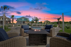A patio area with wicker furniture and a fire pit, overlooking a pool and a building with a balcony.