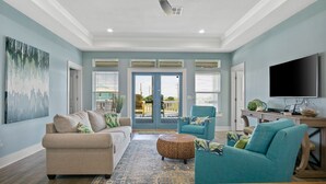 A spacious living room with a beige sofa, teal armchairs, and a wooden coffee table. A large TV is mounted on the wall, and a painting is visible on the left wall. -  Private Heated Pool & Hot Tub ~ Ocean View ~ King Beds ~ Quiet & Peaceful (Bolivar Peninsula)