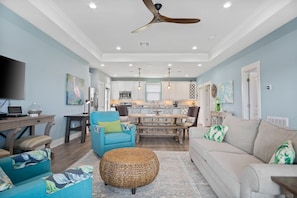 A spacious living room with a large sofa, a blue armchair, and a wicker ottoman. A dining area with a wooden table and chairs is visible in the background.
