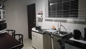 A compact kitchen with a sink, a microwave, and a small refrigerator. There is a dining table with chairs and a window with blinds.