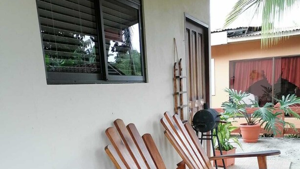 A covered outdoor seating area with wooden chairs and tables, potted plants, and a window with blinds.