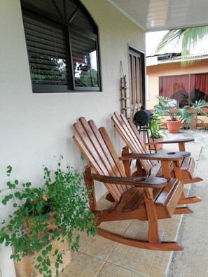 A covered outdoor seating area with wooden chairs and tables, potted plants, and a window with blinds. - A 10 min del Parque de la Fortuna, Apartamento en Zona Tranquila, Parqueo Gratis (La Fortuna)